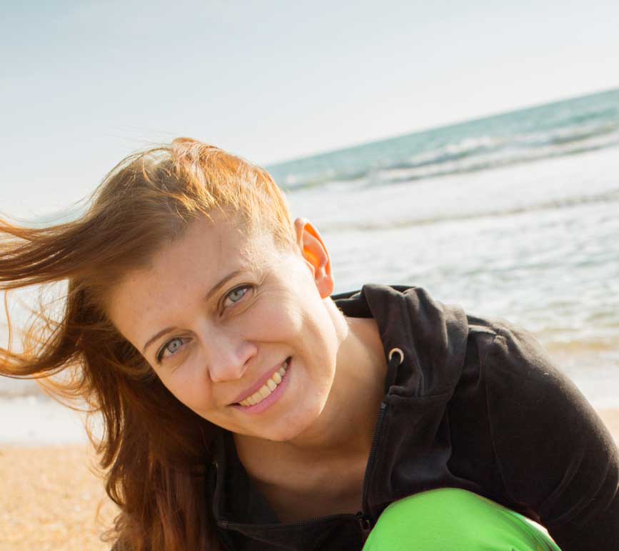Eine Frau mit langen Haaren lächelt an einem sonnigen Strand in die Kamera. Im Hintergrund rollen sanft die Meereswellen und sie trägt einen dunklen Kapuzenpullover. Eine Frau mit langen Haaren lächelt an einem sonnigen Strand in die Kamera. Im Hintergrund rollen sanft die Meereswellen und sie trägt einen dunklen Kapuzenpullover.