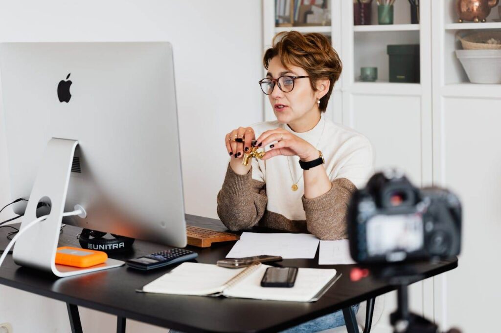 Eine Frau mit Brille sitzt an einem Schreibtisch vor einem großen Computermonitor und hält einen Stift in der Hand. Eine Kamera, ein Smartphone und ein Notebook liegen ebenfalls auf dem Schreibtisch. Im Hintergrund stehen Bücher und Dekoartikel auf Regalen. A professional woman engaged in a virtual meeting setup at her home desk, using a computer and camera.