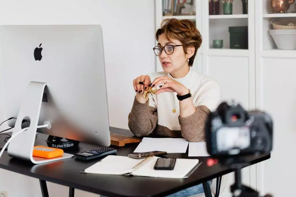Eine Frau mit Brille sitzt an einem Schreibtisch vor einem großen Computermonitor und hält einen Stift in der Hand. Eine Kamera, ein Smartphone und ein Notebook liegen ebenfalls auf dem Schreibtisch. Im Hintergrund stehen Bücher und Dekoartikel auf Regalen. A professional woman engaged in a virtual meeting setup at her home desk, using a computer and camera.