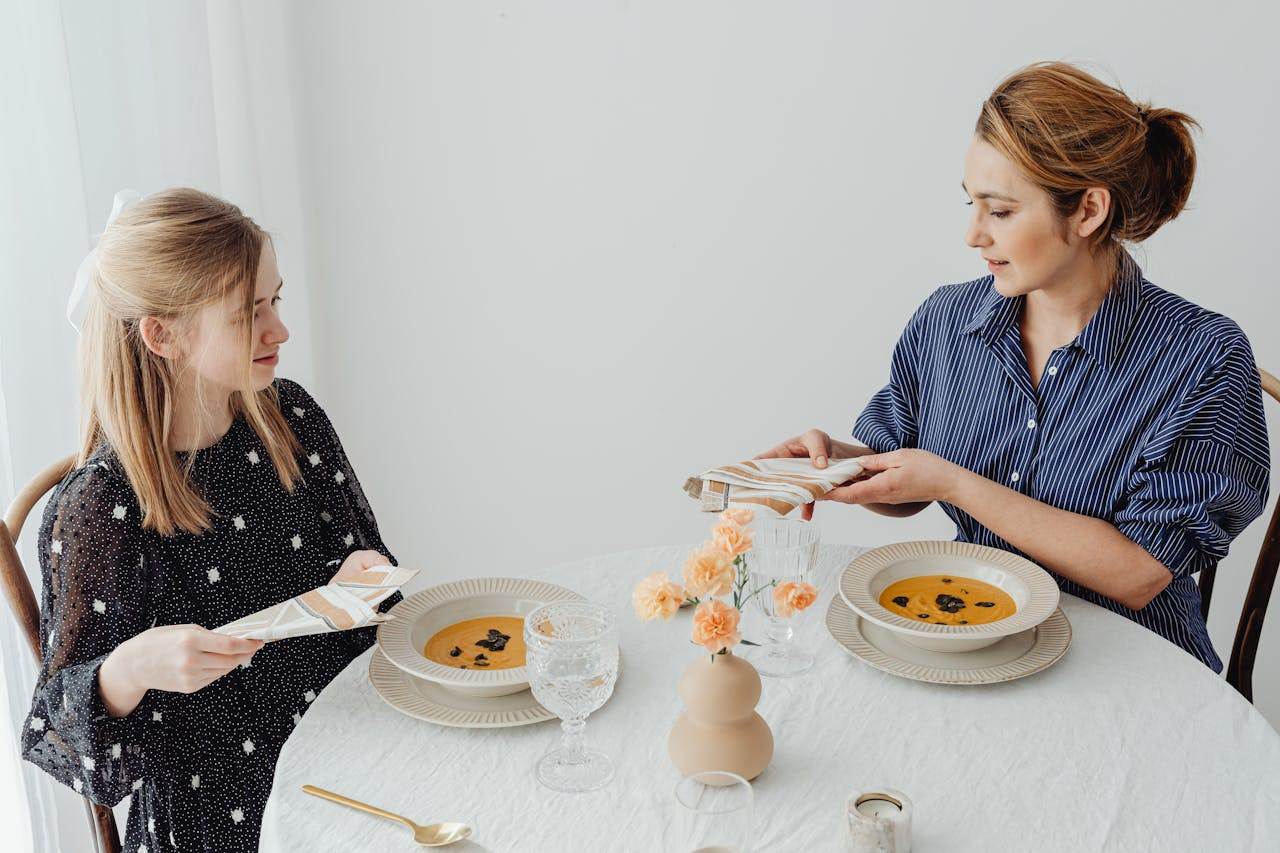 how-we-are Two women dining with soup at an elegantly set table.